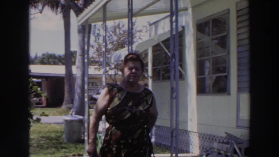 The woman reaches the foreground beside the mobile home, with porch framing, windows, palm trees, lawn, and a blue car still visible behind her.