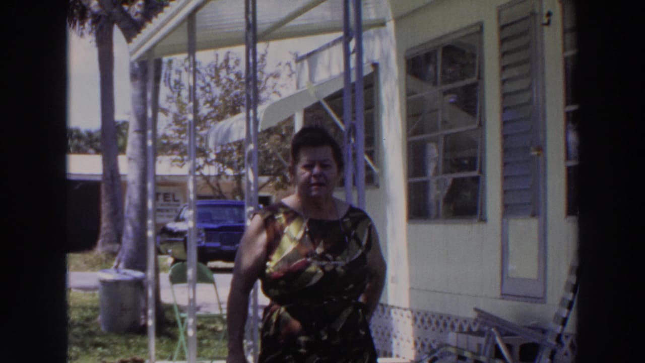 In 1966 Puerto Rico home-movie footage, a woman in a patterned dress faces the camera near a screened porch, with a blue car and palms behind her.