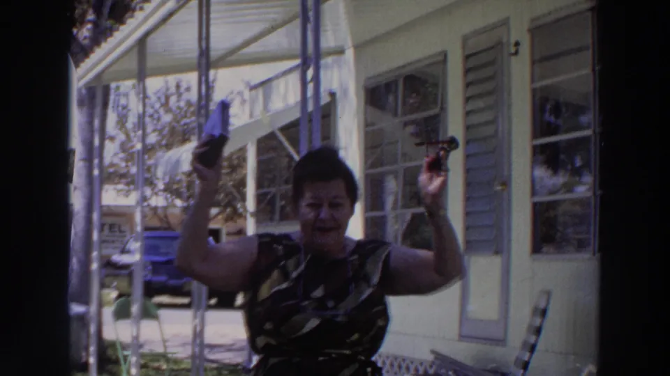 The woman smiles and lifts her handbag and sunglasses beside a screened porch in the 1966 Puerto Rico home-movie scene.