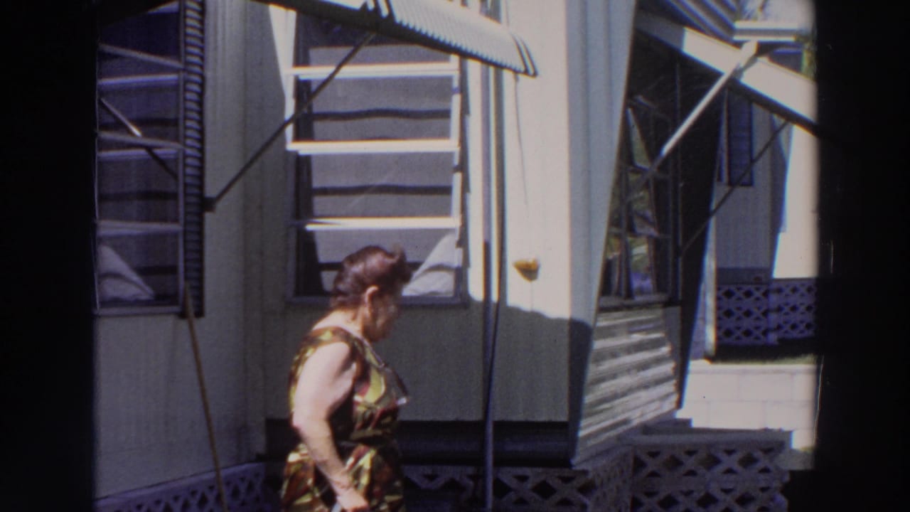 The woman turns and walks away along the porch exterior, with awnings, screen panels, lattice, and steps visible in the 1966 Puerto Rico footage.