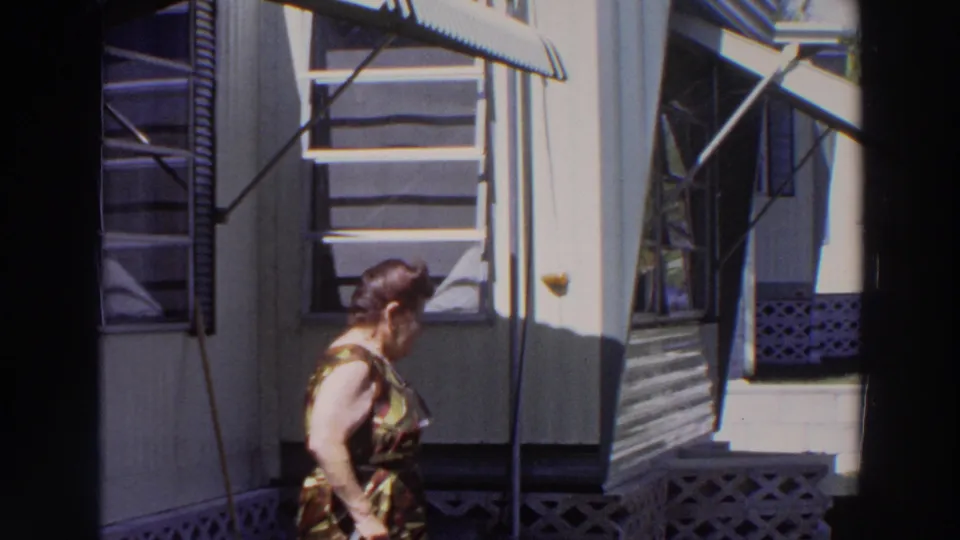 The woman turns and walks away along the porch exterior, with awnings, screen panels, lattice, and steps visible in the 1966 Puerto Rico footage.