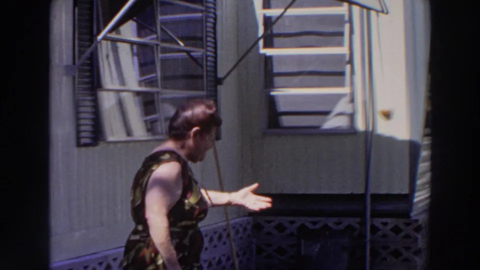 Older woman in a patterned dress gestures animatedly beside a mobile home exterior in Puerto Rico home-movie footage from 1966.