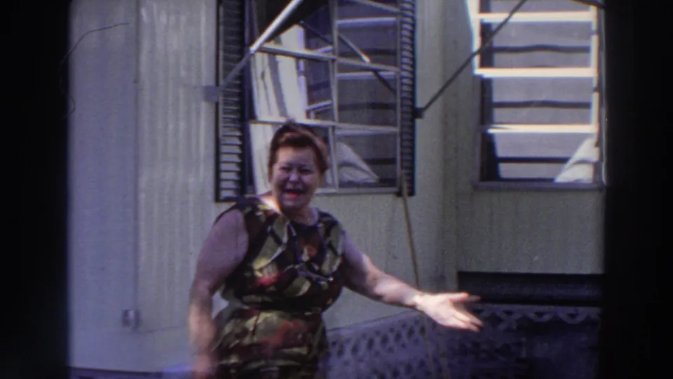 Smiling older woman faces the camera with an arm extended near a light-colored mobile home in 1966 Puerto Rico footage.