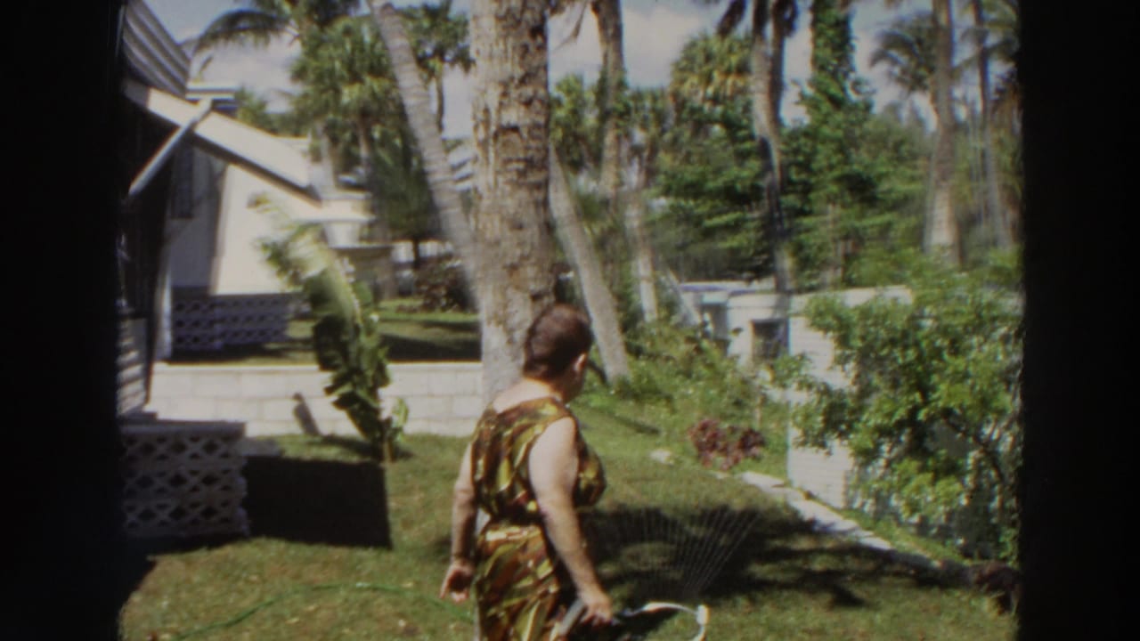 Woman in patterned dress walks across a sunny tropical yard beside a white mobile home.