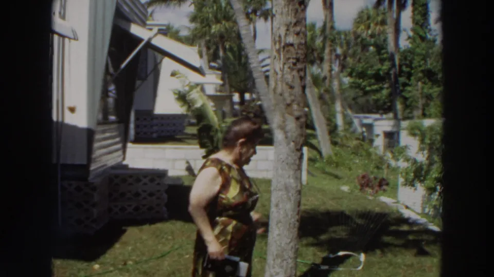 The woman moves past a palm tree in a Puerto Rico yard, with the white mobile home and tropical landscaping behind her.
