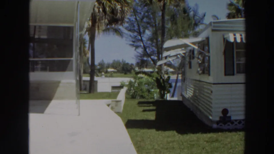 A 1966 Puerto Rico yard view shows a white mobile home with awnings, lawn, palms, walkway, and a bright background beyond the property.