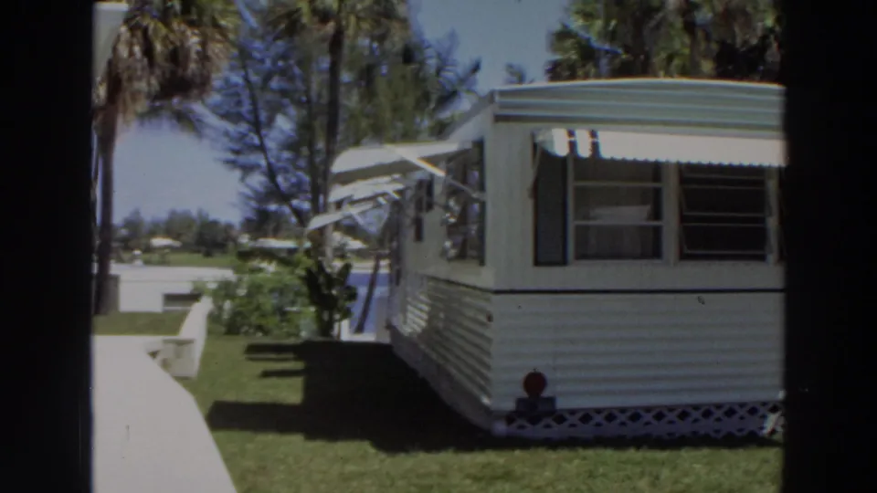 The frame shows the mobile home exterior in Puerto Rico, including awnings, lawn, palm trees, shrubs, skirting, and strong sunlight.