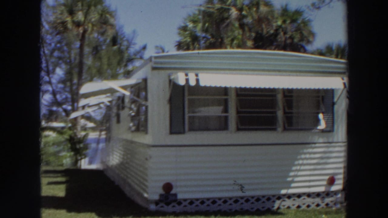 1966 Puerto Rico 8mm frame showing a white mobile home with striped awnings, dark shutters, lattice skirting, grass, and palms.