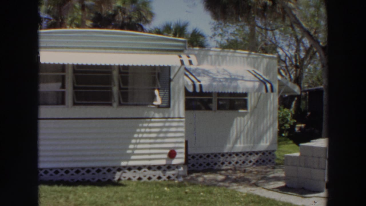 1966 Puerto Rico home-movie frame showing the mobile home from a closer side angle with awnings, windows, lattice skirting, and lawn.