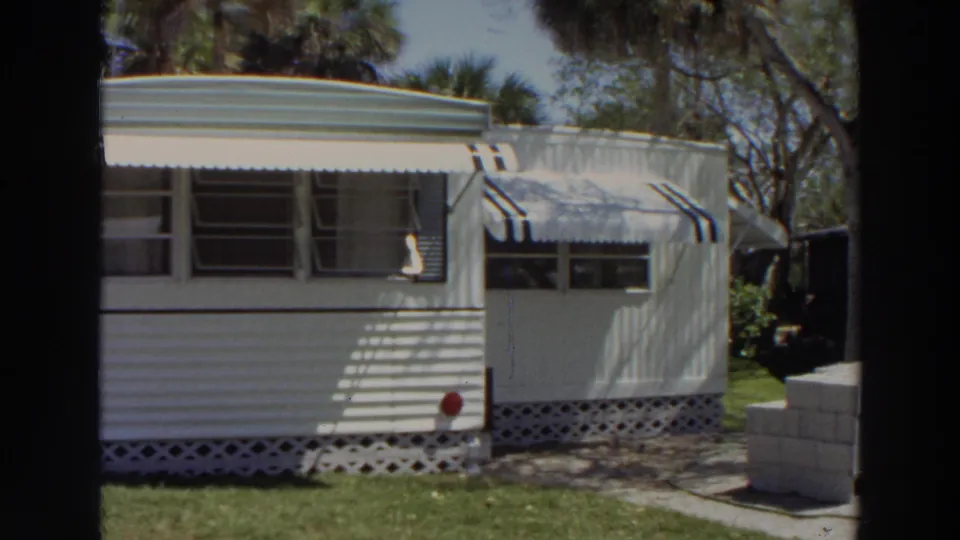 1966 Puerto Rico home-movie frame showing the mobile home from a closer side angle with awnings, windows, lattice skirting, and lawn.
