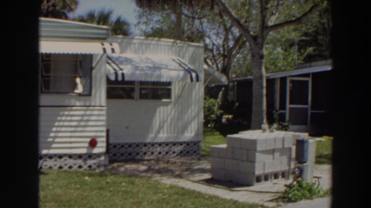 1966 Puerto Rico frame showing the mobile home end, striped awning, trees, lawn, stacked cinder blocks, and a neighboring structure.