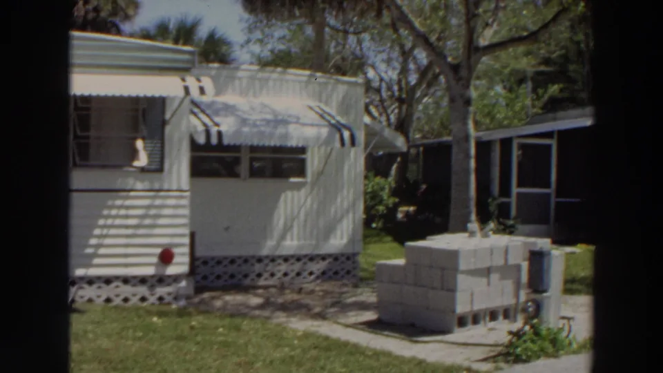 1966 Puerto Rico frame showing the mobile home end, striped awning, trees, lawn, stacked cinder blocks, and a neighboring structure.
