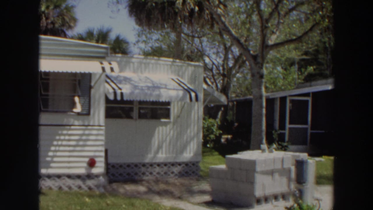 White mobile home with striped awnings, palm trees, grass, and cinder blocks nearby.