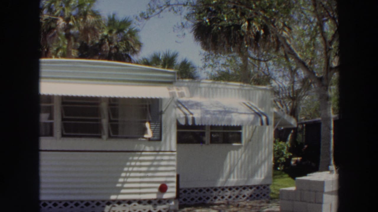 1966 Puerto Rico frame of a white trailer home with awnings, open windows, palm trees, and shaded grass.