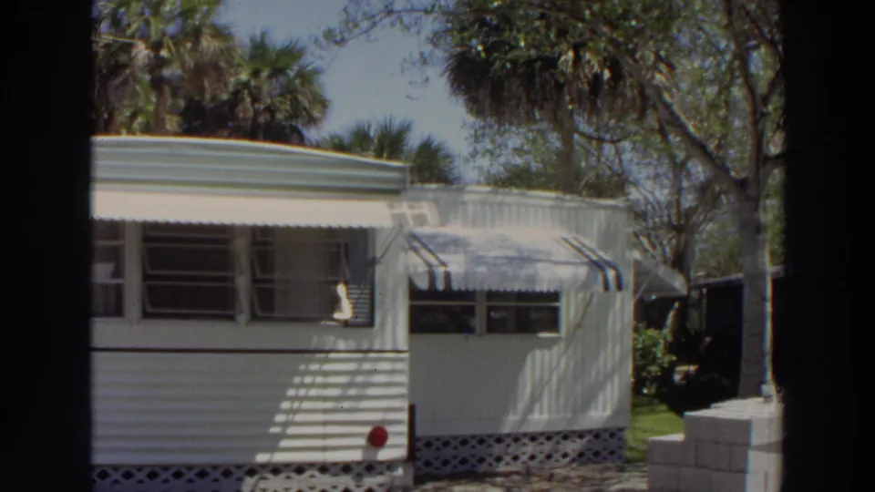 1966 Puerto Rico frame of a white trailer home with awnings, open windows, palm trees, and shaded grass.
