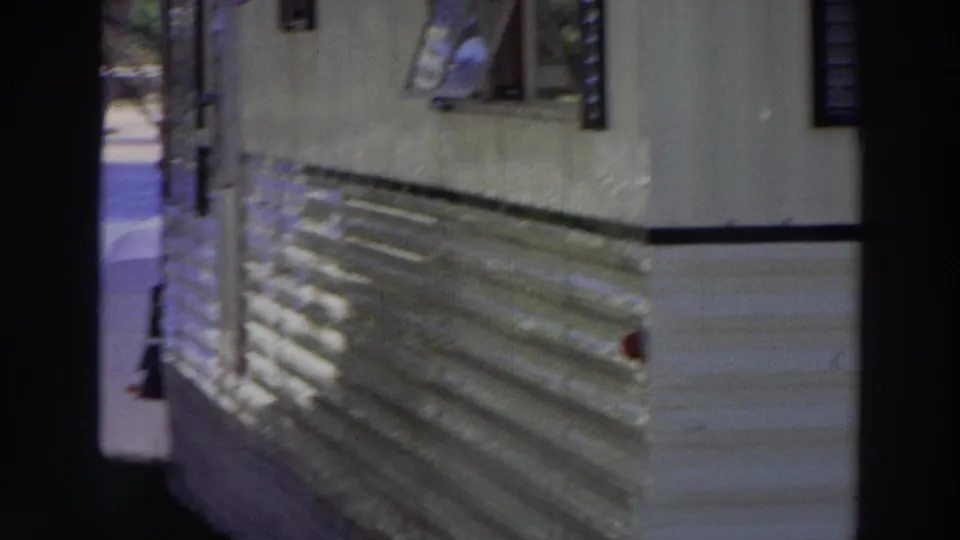 1966 Puerto Rico frame showing the side of a mobile home, small windows, striped awning, and dark porch framing in foreground.