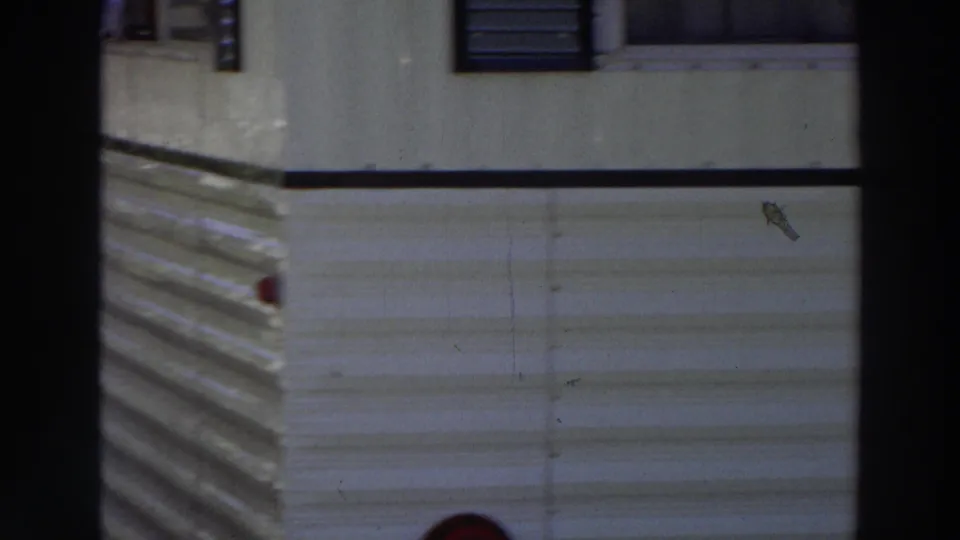 1966 Puerto Rico home-movie frame showing white corrugated mobile-home siding, dark window bars, and a small red exterior light.