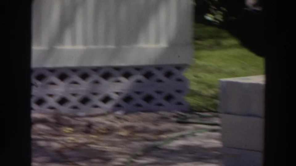 1966 Puerto Rico frame showing the mobile home's lower siding, lattice foundation, grass, stone edging, and nearby concrete blocks.