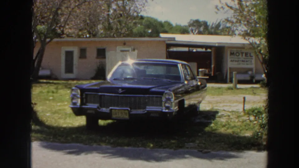 A dark blue American sedan sits on grass outside a Puerto Rico roadside motel, with a white motel-apartments sign in the background.