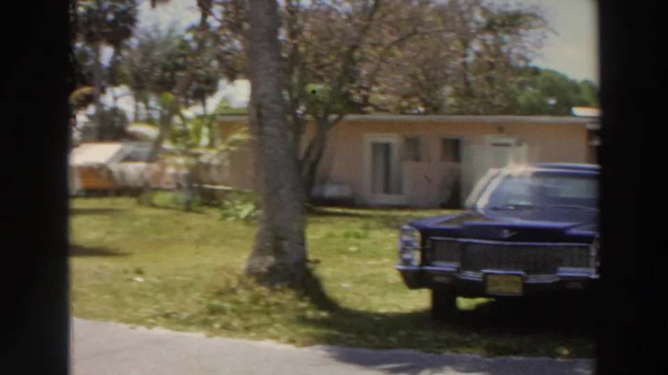 The parked dark blue sedan is seen again on the motel lawn in Puerto Rico, partly cropped by a tree and handheld film framing.
