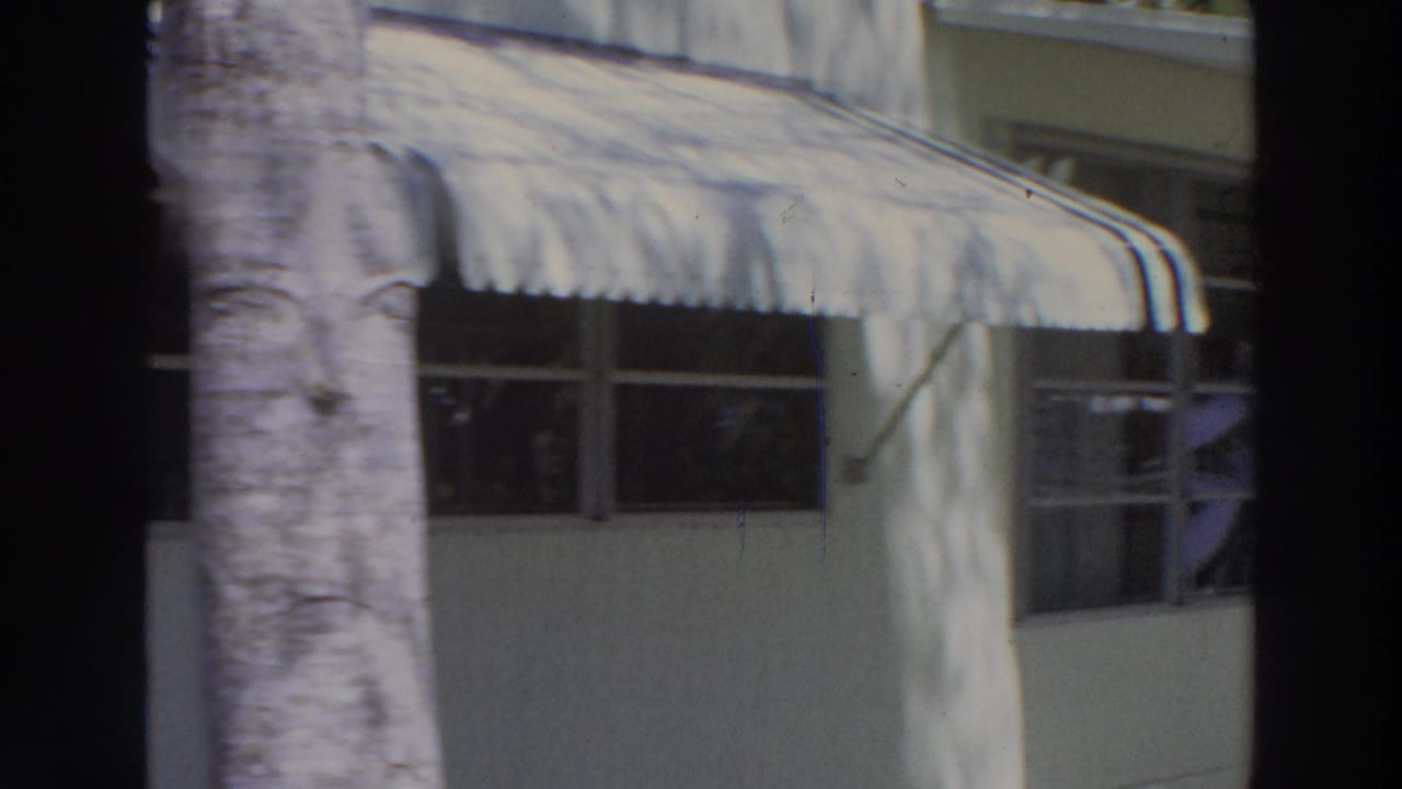 1966 Puerto Rico home-movie frame showing a white stucco house facade with shaded windows, striped awning, palm trunk, and dappled light.