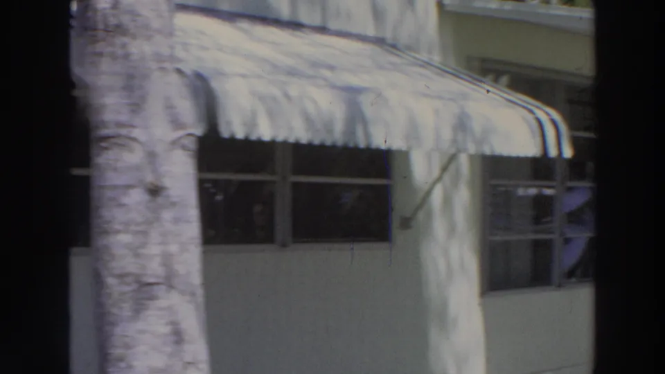 1966 Puerto Rico home-movie frame showing a white stucco house facade with shaded windows, striped awning, palm trunk, and dappled light.