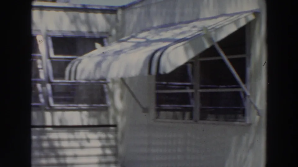 1966 Puerto Rico frame with striped window awnings, metal supports, white stucco surfaces, and dappled tree shadows across the facade.