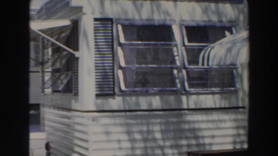 1966 Puerto Rico home-movie frame showing a white facade with louvered panel, projecting window awnings, palm trunk, and strong foliage shadows.