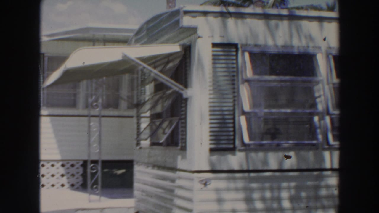 1966 Puerto Rico home-movie frame showing a white mobile home exterior with awning, jalousie windows, lattice skirting, and bright sun.