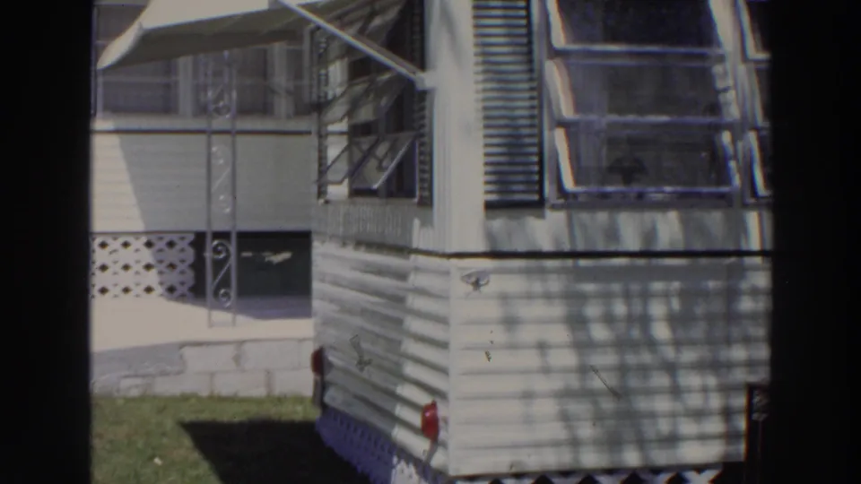 1966 Puerto Rico frame of a resort mobile home corner with white siding, shaded windows, awnings, and grass at the base.