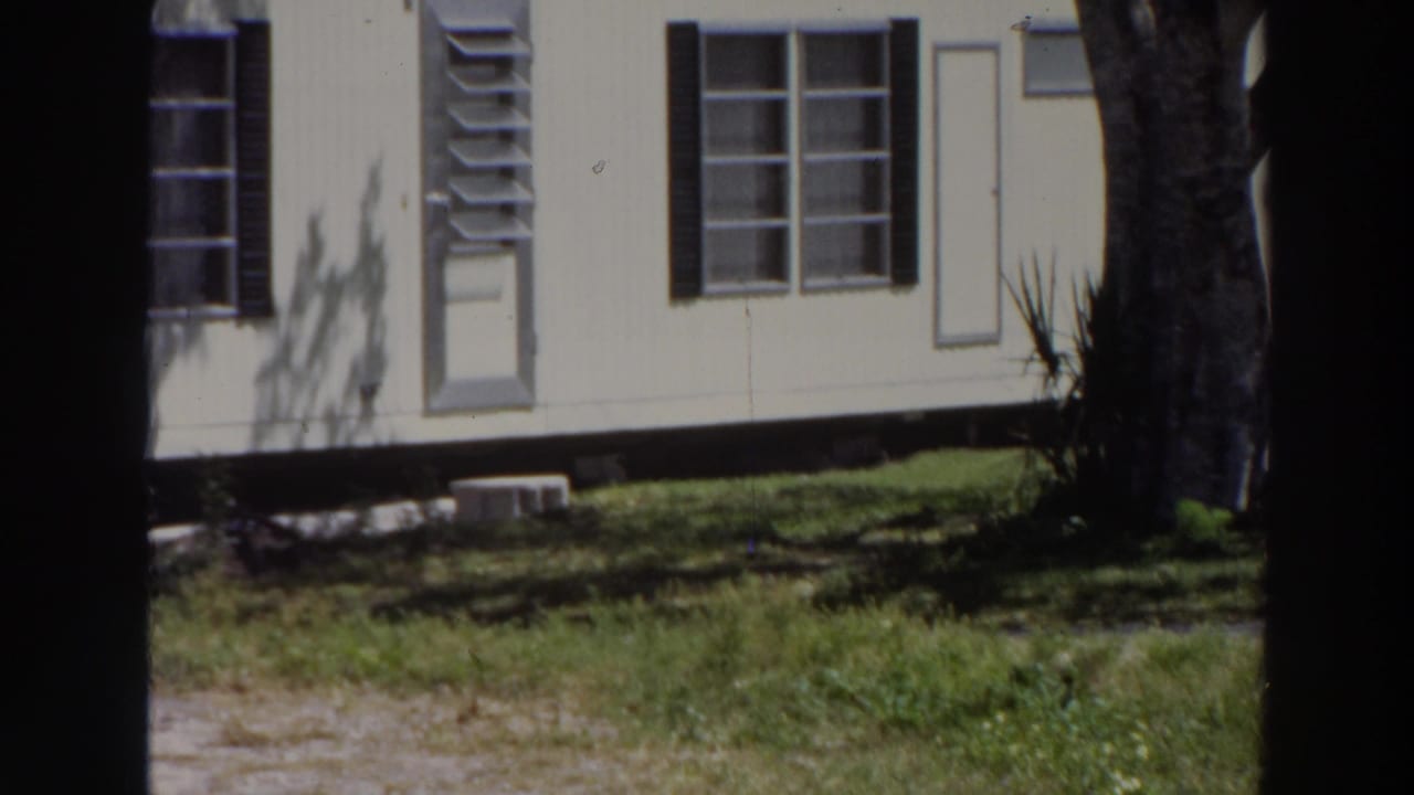 1966 Puerto Rico frame showing the side of a white mobile home with shuttered windows, grass, a tree trunk, and deep afternoon shadows.