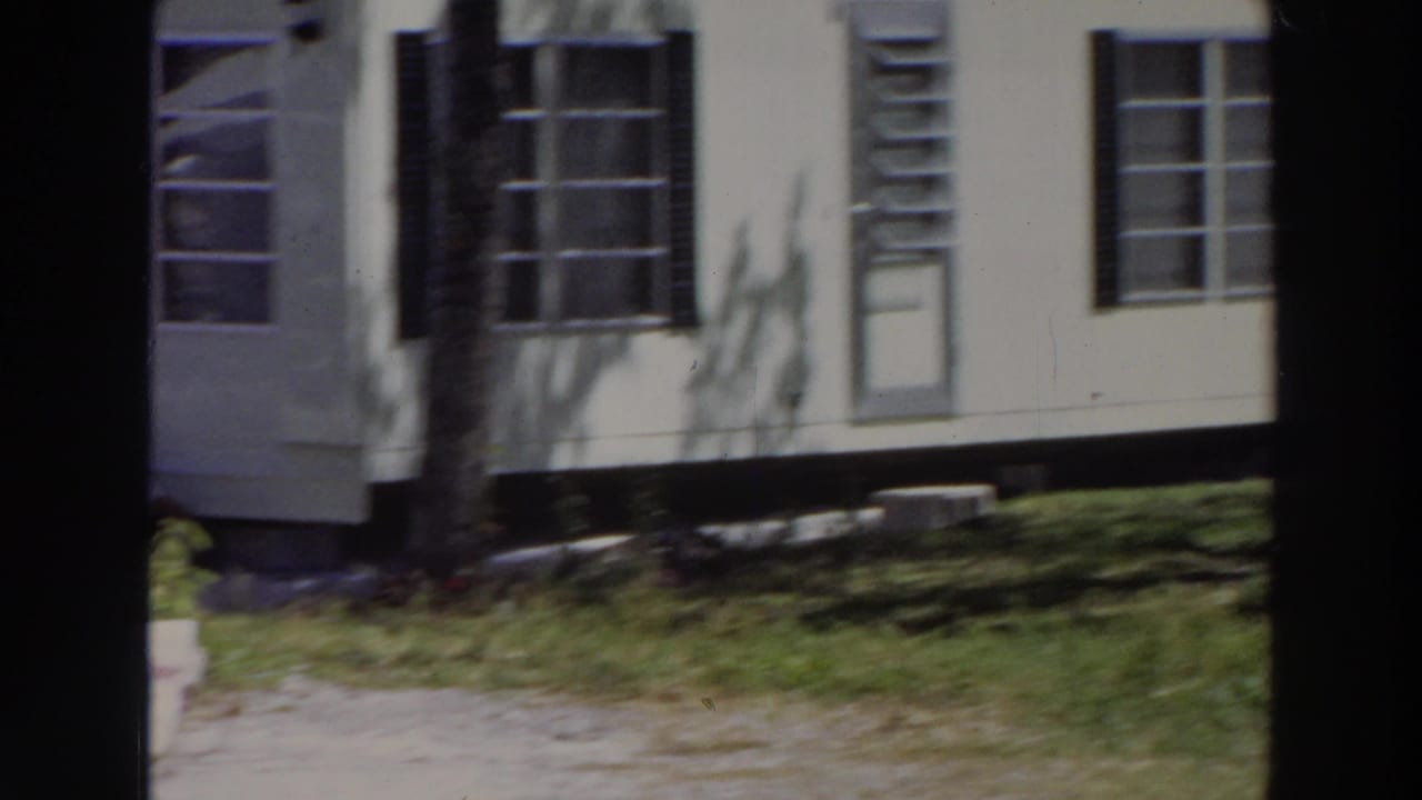 White mobile home exterior with black shutters, windows, grass, and a tree trunk at left.