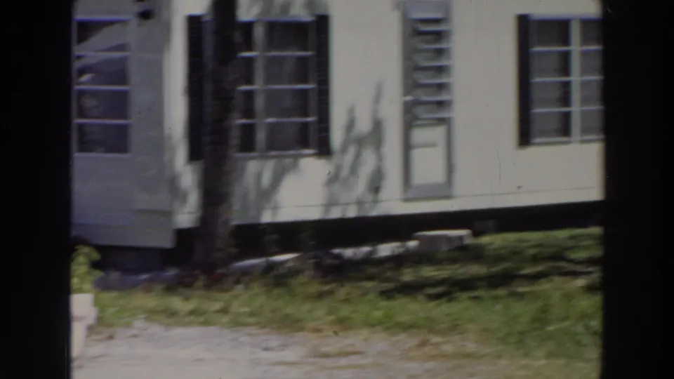 1966 Puerto Rico home-movie frame showing a white mobile home with black shutters, multi-pane windows, louvered vents, grass, and a tree trunk.