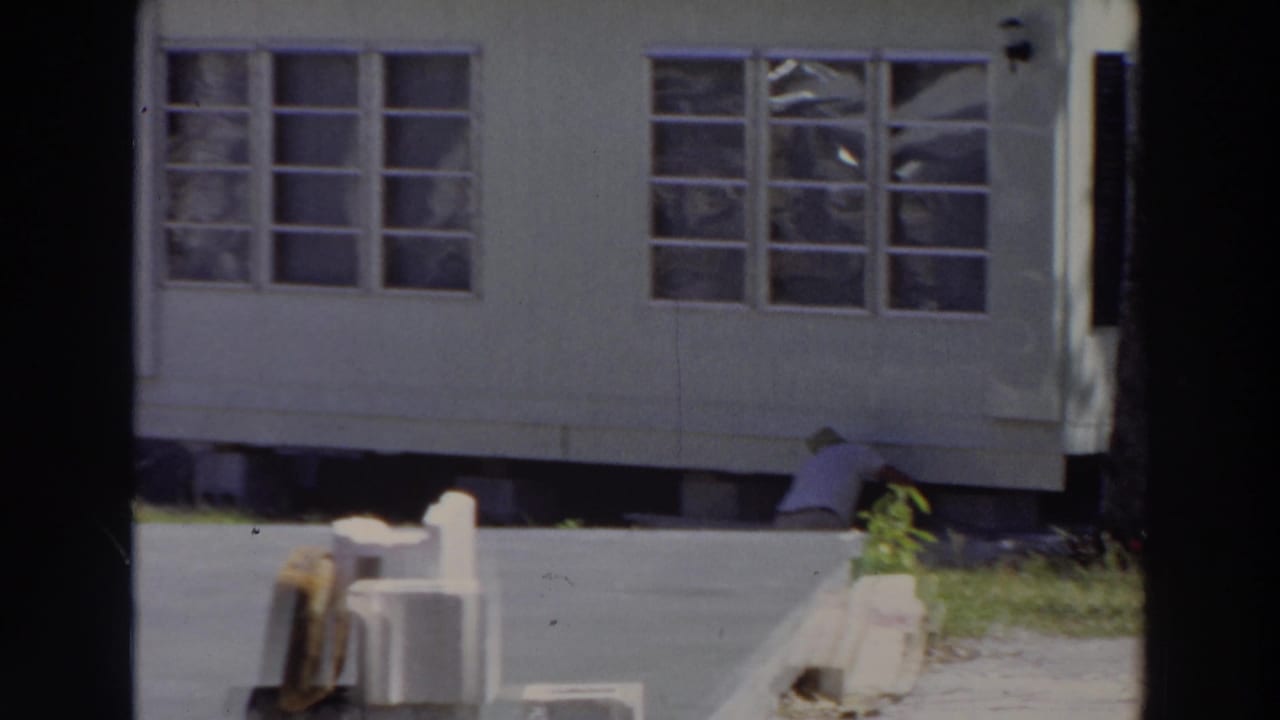 White mobile home with rows of windows, paved ground, and a crouching person near the foundation.