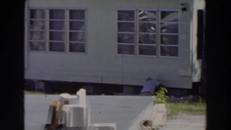 1966 Puerto Rico frame showing a white mobile home, wide windows, paved ground, and a person crouched near the raised foundation.