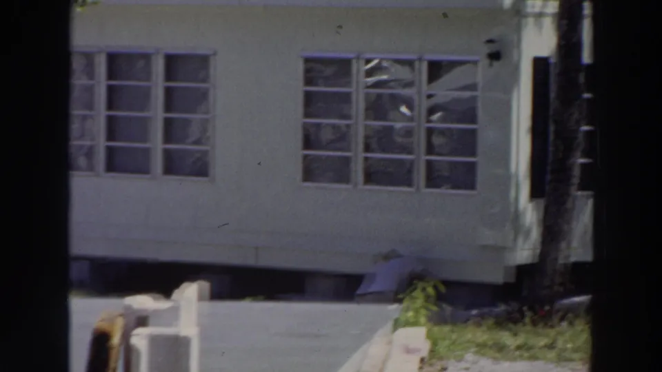 1966 Puerto Rico home-movie frame with a white mobile home, rectangular windows, paved ground, grass, and a partially visible person near the base.