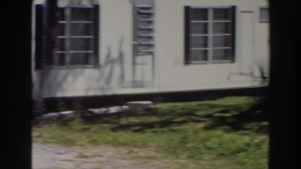 1966 Puerto Rico frame showing the side of a white mobile home with black shutters, louvered vent, windows, grass, and tree shadows.