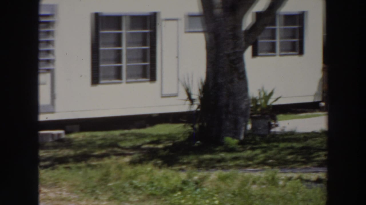 1966 Puerto Rico 8mm frame showing a light house facade with dark shutters, multi-pane windows, tree trunk, and shaded lawn.