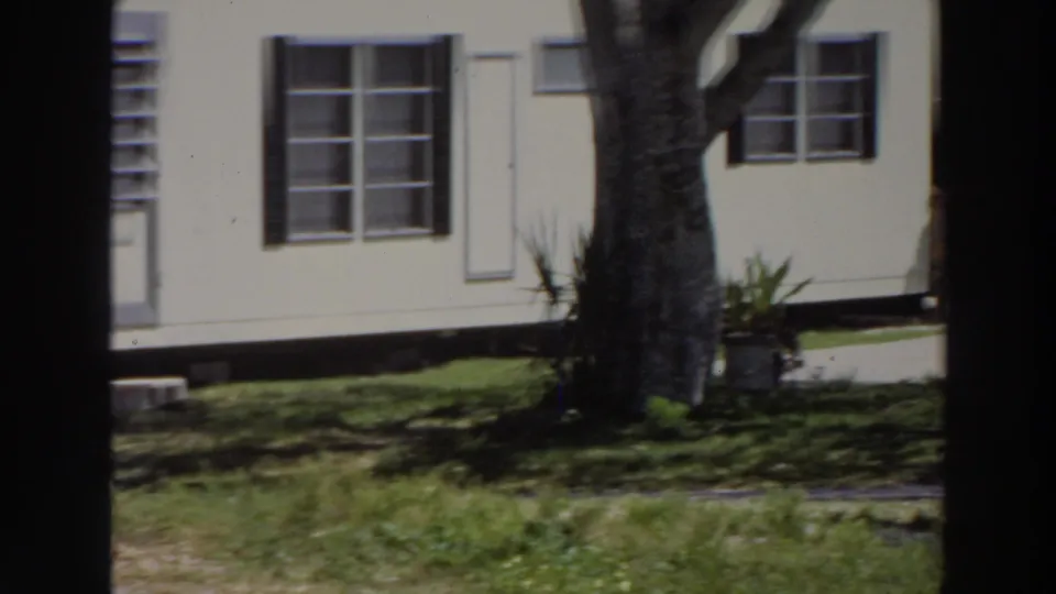 1966 Puerto Rico 8mm frame showing a light house facade with dark shutters, multi-pane windows, tree trunk, and shaded lawn.