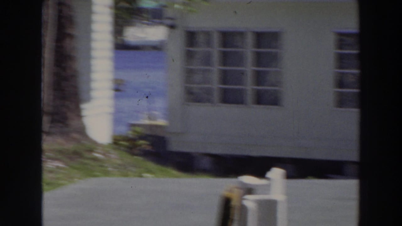 1966 Puerto Rico frame showing the corner of a light-colored home, multi-pane windows, lawn, paved drive, tree trunk, and short posts.