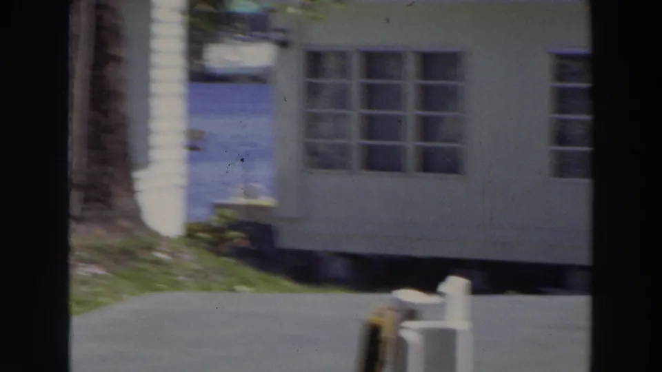 1966 Puerto Rico frame showing the corner of a light-colored home, multi-pane windows, lawn, paved drive, tree trunk, and short posts.