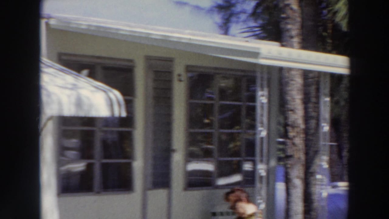 1966 Puerto Rico home-movie frame showing a covered porch, awning, windows, tree trunks, and a seated woman partly visible near the lower edge.