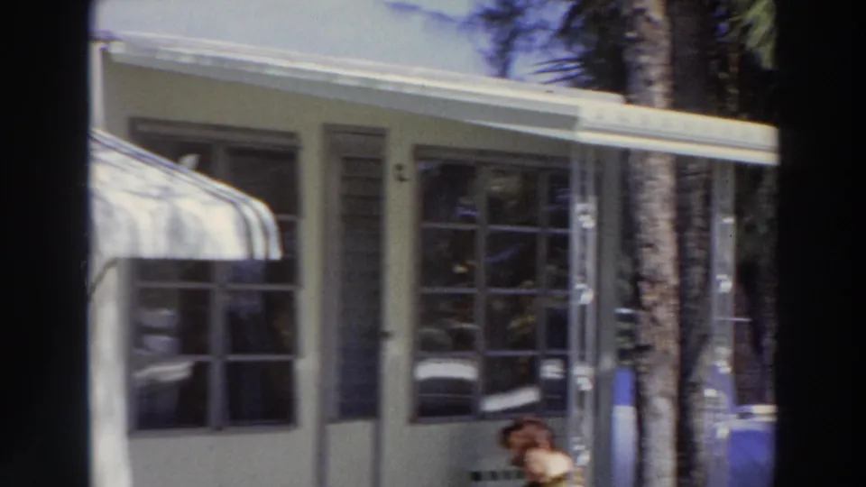 1966 Puerto Rico home-movie frame showing a covered porch, awning, windows, tree trunks, and a seated woman partly visible near the lower edge.