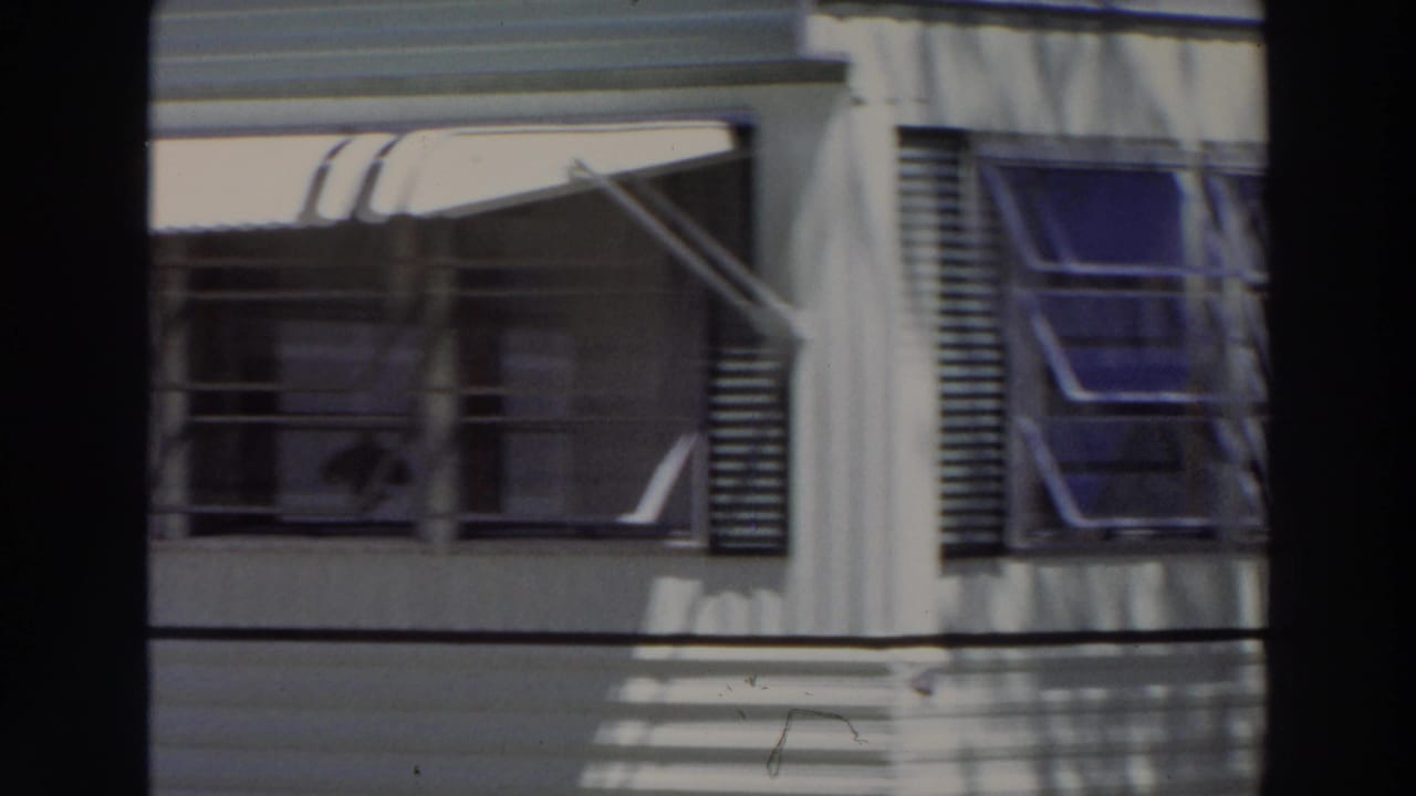 1966 Puerto Rico frame showing windows, open louvered shutters, awning supports, and dark palm shadows on a pale facade.