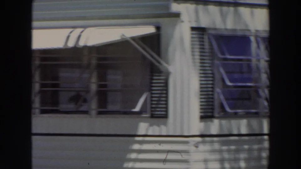 1966 Puerto Rico frame showing windows, open louvered shutters, awning supports, and dark palm shadows on a pale facade.