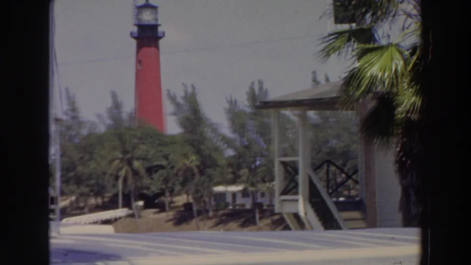 1966 Puerto Rico home-movie frame showing a red lighthouse above foliage, with palm trees, a small structure, and paved foreground.