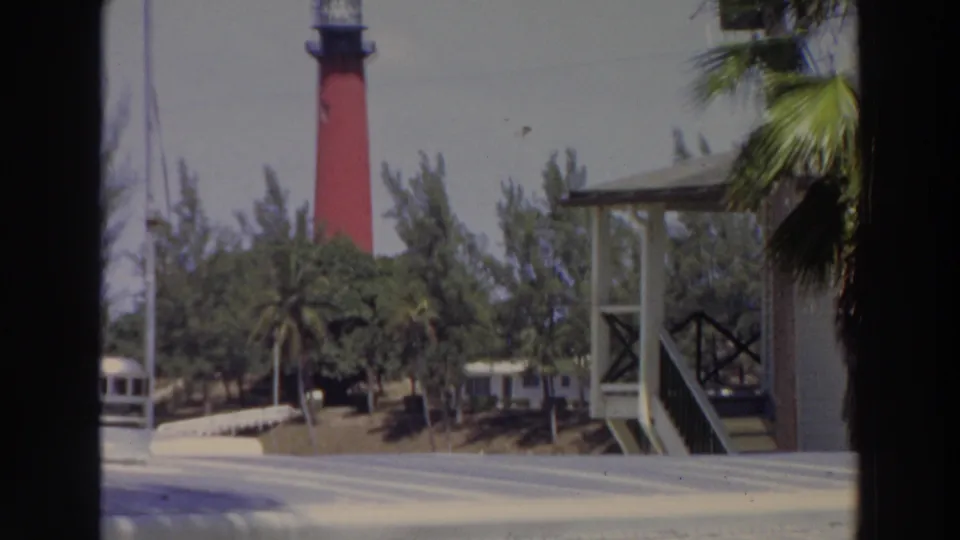 1966 Puerto Rico frame with a red lighthouse partly framed by palms, dense trees, a small structure, and bright paved foreground.