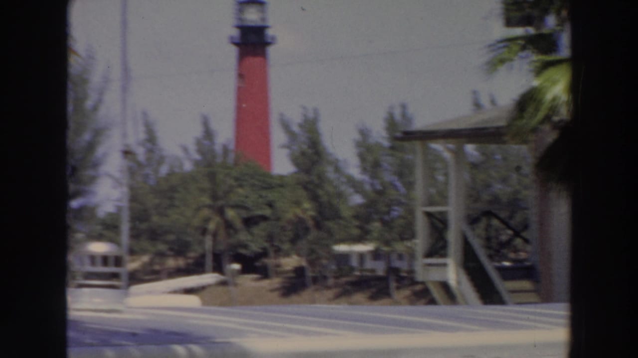 Red lighthouse behind palms and foliage, seen through dark vertical frame edges.