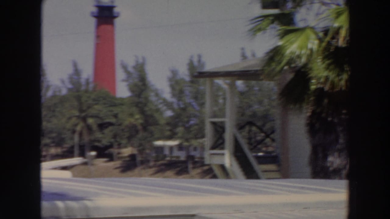 Red lighthouse and white porch structure framed by palms and dark vertical bars.