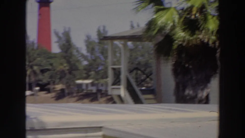 1966 Puerto Rico home-movie frame with a red lighthouse partly obscured by palms, a porch structure, and road surface in front.